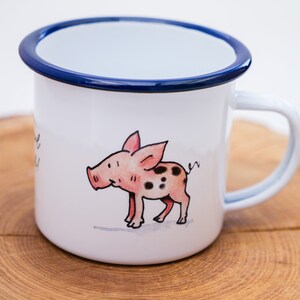 May include: White enamel mug with a blue rim and a hand-painted illustration of a pink pig with black spots. The pig is looking to the left and has a curly tail.