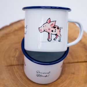 May include: Two white enamel mugs with blue rims. The top mug features a cartoon pig with pink spots. The bottom mug has the text "Sassi Girl Gl&uuml;ck!" in black.