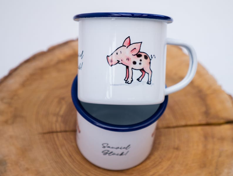 May include: Two white enamel mugs with blue rims. The top mug features a cartoon pig with pink spots. The bottom mug has the text "Sassi Girl Gl&uuml;ck!" in black.