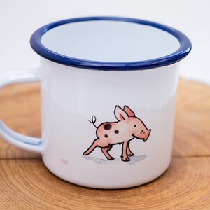 May include: White enamel mug with a blue rim and a hand-drawn illustration of a pink pig with black spots.