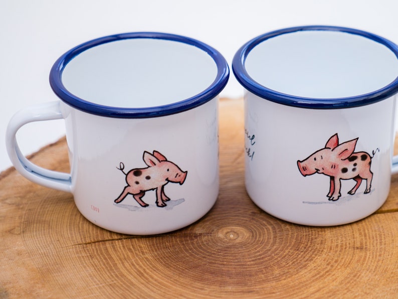 May include: Two white enamel mugs with blue rims. Each mug features a cartoon illustration of a pink pig with black spots. One mug has the text "You are my sunshine" on it.
