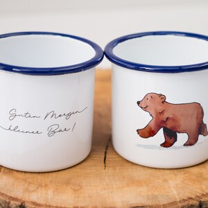 May include: Two white enamel mugs with blue rims. One mug has a brown bear illustration and the other has the text "Guten Morgen kleiner B&auml;r!" with a red robin illustration.
