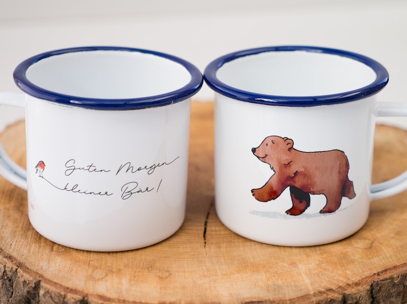 May include: Two white enamel mugs with blue rims. One mug has a brown bear illustration and the other has the text "Guten Morgen kleiner B&auml;r!" with a red robin illustration.