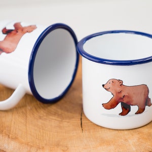 May include: Two white enamel mugs with blue rims. One mug features a brown bear walking to the right. The other mug features a brown bear walking to the left.