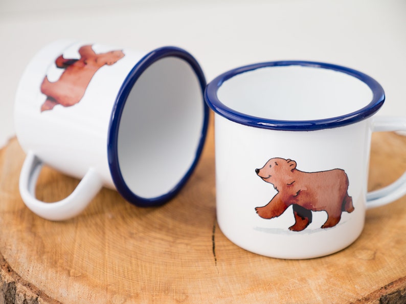 May include: Two white enamel mugs with blue rims. One mug features a brown bear walking to the right. The other mug features a brown bear walking to the left.