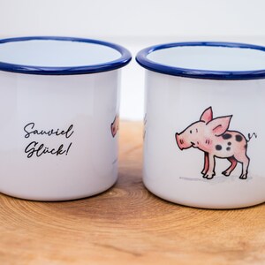 May include: Two white enamel mugs with blue rims. One mug features a cartoon pig with black spots and the text "Sawviel Gl&uuml;ck!" The other mug features a cartoon pig with black spots.
