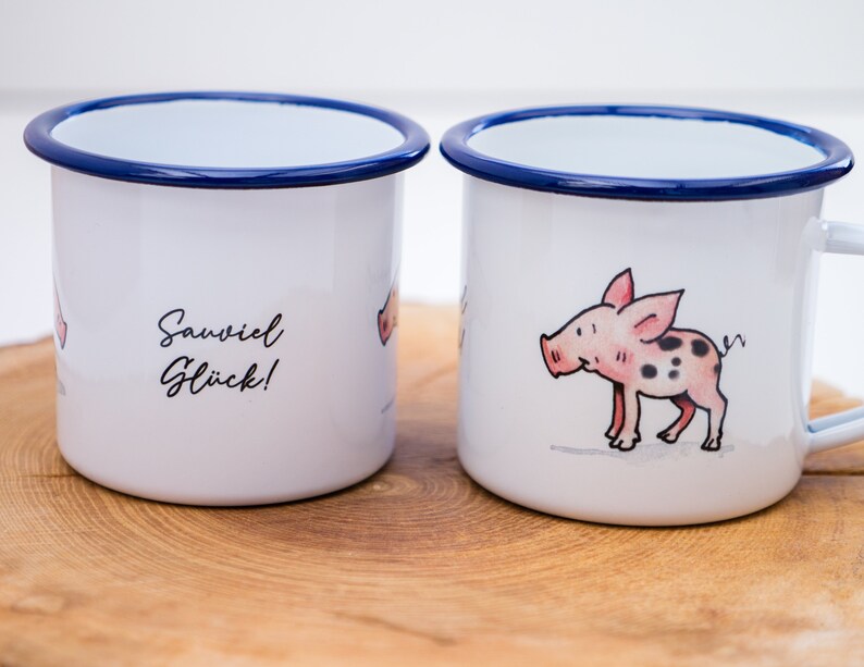 May include: Two white enamel mugs with blue rims. One mug features a cartoon pig with black spots and the text "Sawviel Gl&uuml;ck!" The other mug features a cartoon pig with black spots.