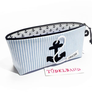 Könnte beinhalten: Eine blau-weiß gestreifte Kosmetiktasche mit einem schwarzen Anker-Applikation und einem weißen Seildetail. Die Tasche hat einen schwarzen Reißverschluss und ein weißes Innenfutter mit kleinen schwarzen Anker-Mustern. Das Wort "FRIEDA" steht auf einem kleinen Etikett.