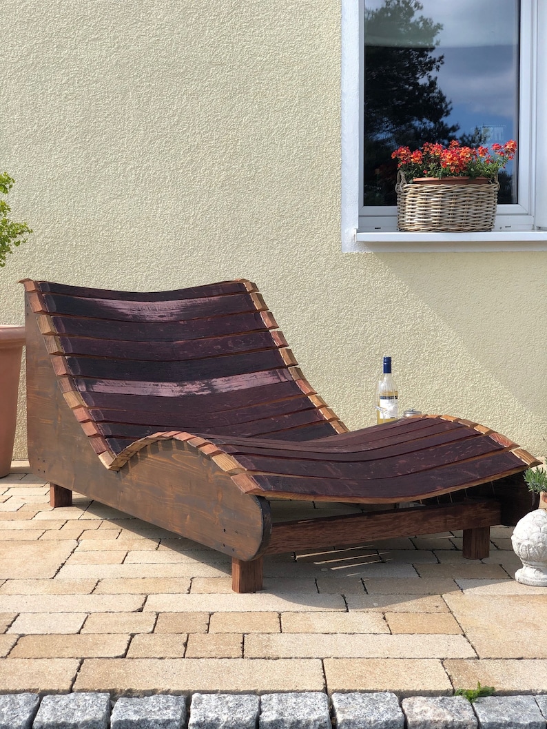 K&ouml;nnte beinhalten: Eine Chaiselongue aus Holz, hergestellt aus wiederverwendeten Weinf&auml;ssern. Die Liege ist dunkelbraun mit einem geschwungenen Design und steht auf einer Terrasse.