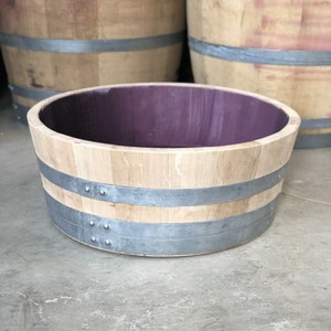 Könnte beinhalten: Ein kleiner Holzfass-Pflanzer mit zwei Metallbändern und einem violetten Innenraum. Der Pflanzer steht auf einer Betonfläche.