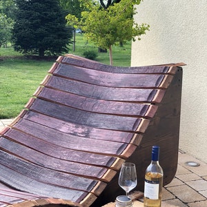 K&ouml;nnte beinhalten: Ein Holz-Liegestuhl aus wiederverwendeten Weinfassdauben. Der Stuhl ist gebogen und hat eine nat&uuml;rliche Holzoptik. Vor dem Stuhl steht eine kleine Holzkiste mit einer Flasche Wei&szlig;wein und einem Glas.