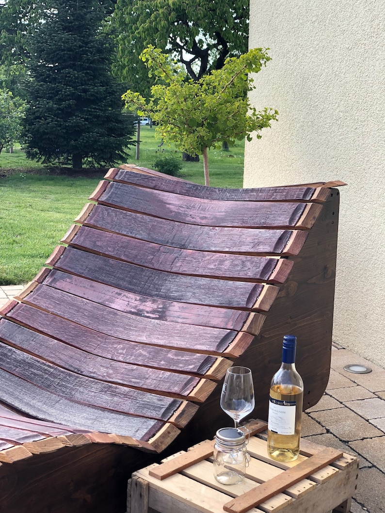 K&ouml;nnte beinhalten: Ein Holz-Liegestuhl aus wiederverwendeten Weinfassdauben. Der Stuhl ist gebogen und hat eine nat&uuml;rliche Holzoptik. Vor dem Stuhl steht eine kleine Holzkiste mit einer Flasche Wei&szlig;wein und einem Glas.