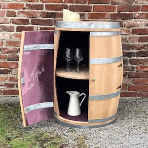 May include: A wooden wine barrel repurposed as a cabinet with a hinged door, two shelves, and a white pitcher inside. The cabinet is made of light brown wood with silver metal accents. The door is open and reveals a purple interior.