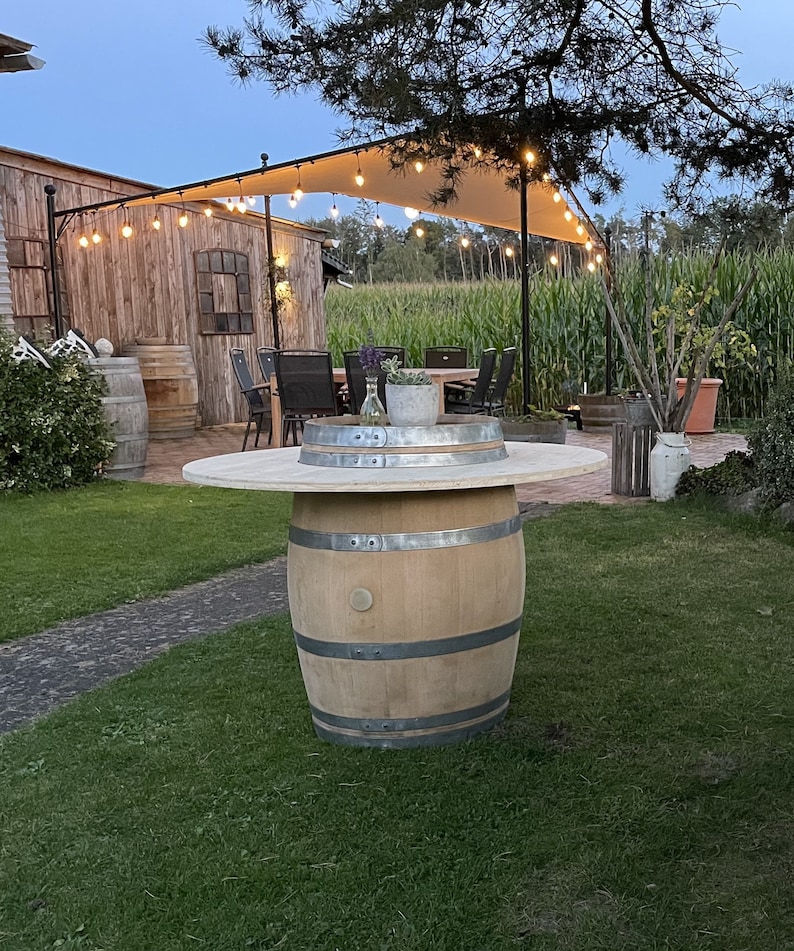 May include: A wooden barrel table with a white tabletop, surrounded by green grass and a wooden building in the background. The table is set up for outdoor dining with string lights overhead.