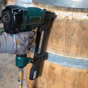 May include: A green and black pneumatic nail gun is being used to attach a metal band to a wooden barrel. The nail gun has a hose attached to it and the text "PREZENA AC-ZS0" is visible on the side.