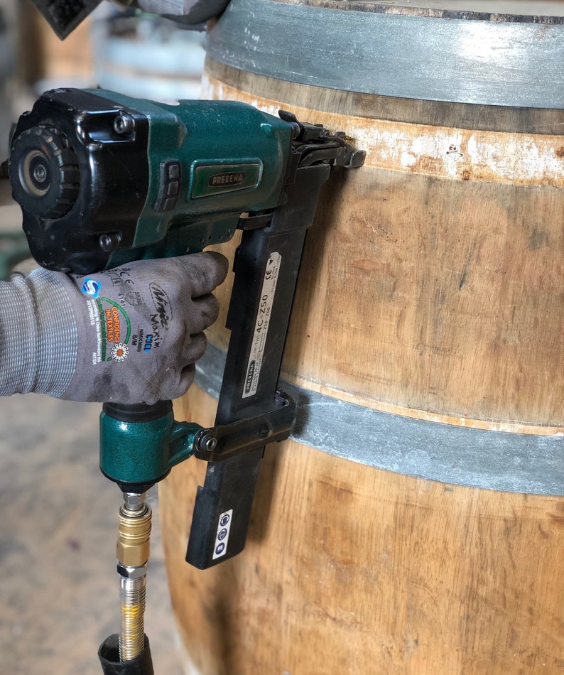 May include: A green and black pneumatic nail gun is being used to attach a metal band to a wooden barrel. The nail gun has a hose attached to it and the text "PREZENA AC-ZS0" is visible on the side.
