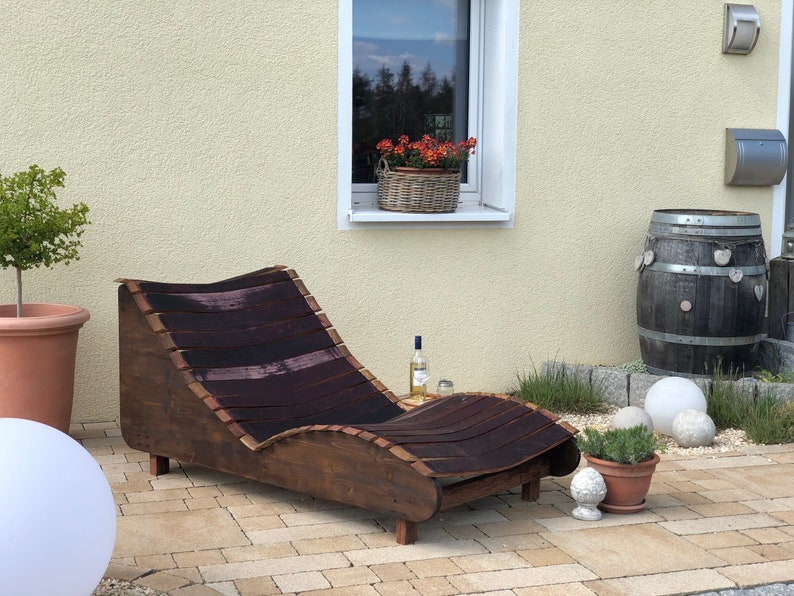 K&ouml;nnte beinhalten: Ein Holz-Liegestuhl aus wiederverwendeten Weinf&auml;ssern. Der Stuhl ist braun mit einem dunkelbraunen Streifenmuster und steht auf einer Terrasse mit einem Fass und Topfpflanzen.