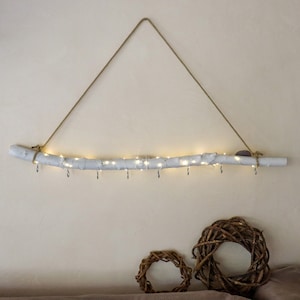 Könnte beinhalten: Eine weiß lackierte Treibholz-Wanddekoration mit kleinen silbernen Haken und einer Lichterkette mit warmweißen Feenlichtern. Das Treibholz wird von einem Naturseil gehalten. Zwei kleine, runde, geflochtene Kränze befinden sich darunter.