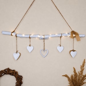 Könnte beinhalten: Ein weiß bemalter Holzzweig als Wandbehang, verziert mit warmweißen Lichterketten und fünf hängenden weißen Herzornamenten an Bindfäden. Eine beigefarbene Schleife mit Punkten ist rechts am Zweig befestigt, der an einer braunen Kordel hängt.