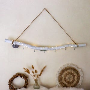 Könnte beinhalten: Weiß lackierter Ast als Wandbehang mit kleinen Haken und Lichterketten. Der Ast wird von einem braunen Seil gehalten und hat kleine Metallhaken zum Aufhängen von Gegenständen. Dekorative Elemente darunter.