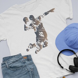 Könnte beinhalten: Weißes T-Shirt mit einer Silhouette einer Person, die einen Ball wirft, gefüllt mit einem Tarnmuster in Braun- und Blautönen. Eine blaue Baseballkappe, Jeans und schwarze Kopfhörer sind ebenfalls zu sehen.