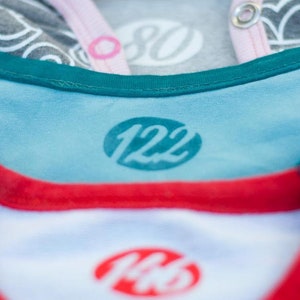 May include: Close-up of three different coloured shirts with size labels. The top shirt is grey with a white label that says "80". The middle shirt is blue with a green label that says "122". The bottom shirt is white with a red label that says "146".