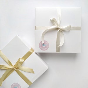 May include: Two white gift boxes with gold and ivory ribbon bows. The boxes are tied with ribbon and have a sticker with a whale design and the text "Little Toys Tatoockairy's".