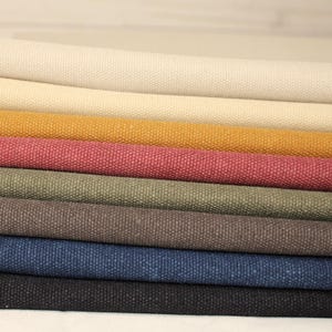 Könnte beinhalten: Nahaufnahme von mehreren Stoffrollen in verschiedenen Farben. Die Farben umfassen Creme, Senfgelb, Staubrosa, Olivgrün, Dunkelbraun, Marineblau und Schwarz. Der Stoff hat ein strukturiertes Gewebe.