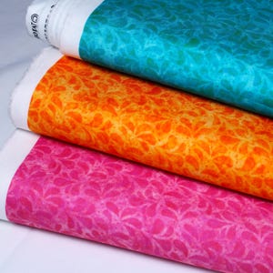 Könnte beinhalten: Drei Stoffrollen in den Farbtönen Türkis, Orange und Pink. Jede Stoffrolle zeigt ein sich wiederholendes Muster aus blattartigen Formen. Der Stoff ist eng gerollt, wobei der weiße Rand des Stoffes sichtbar ist.