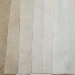 May include: A collection of rectangular fabric swatches in varying shades of beige and off-white. The swatches are arranged vertically, showcasing a gradient from dark to light tones. The edges are raw and unfinished.
