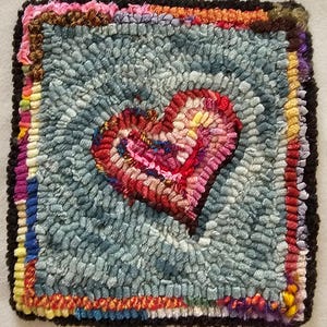 May include: A colorful rug with a heart design in the center. The heart is made of red, pink, and yellow yarn. The rug is surrounded by a border of different colored yarn.