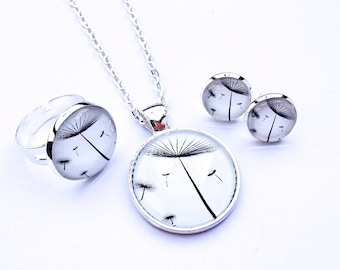 May include: A silver necklace, ring, and earrings set with a dandelion design. The dandelion is black and white and is set in a clear glass circle.