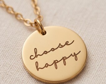 May include: A gold-toned circular pendant necklace featuring the engraved text "choose happy" in a cursive script font. The pendant is suspended from a matching gold-toned metal chain, showcasing a smooth, polished finish on the circular disc.