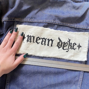 Pode incluir: Uma jaqueta de ganga com um remendo branco costurado nas costas. O remendo tem as palavras "mean dyke" escritas em letras góticas pretas.