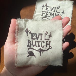 K&ouml;nnte beinhalten: Zwei kleine, quadratische Aufn&auml;her mit rohen Kanten. Auf einem Aufn&auml;her steht "EVIL BUTCH" in dunkelbrauner Tinte mit dekorativen Verzierungen. Der andere Aufn&auml;her tr&auml;gt die Aufschrift "EVIL FEMME" in &auml;hnlichem Stil. Die Aufn&auml;her werden in einer Hand gehalten.