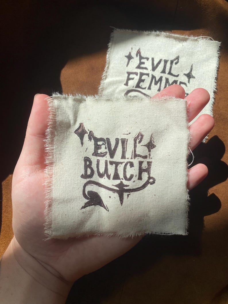 K&ouml;nnte beinhalten: Zwei kleine, quadratische Aufn&auml;her mit rohen Kanten. Auf einem Aufn&auml;her steht "EVIL BUTCH" in dunkelbrauner Tinte mit dekorativen Verzierungen. Der andere Aufn&auml;her tr&auml;gt die Aufschrift "EVIL FEMME" in &auml;hnlichem Stil. Die Aufn&auml;her werden in einer Hand gehalten.