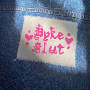 May include: A denim jacket with a white patch featuring pink text and heart graphics. The text appears to read "Smoke Slut". The jacket has a light blue wash and a small pocket.