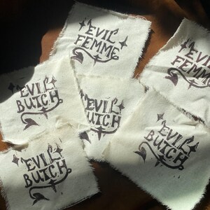 K&ouml;nnte beinhalten: Mehrere cremefarbene Stoffflicken mit den Aufschriften "EVIL FEMME" und "EVIL BUTCH" in dunkelbrauner Tinte. Der Text ist mit dekorativen Elementen versehen. Die Flicken haben rohe, unfertige Kanten und liegen auf einer braunen Oberfl&auml;che.