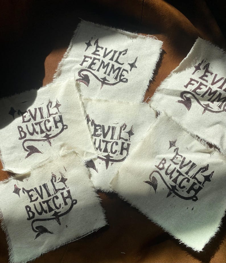 K&ouml;nnte beinhalten: Mehrere cremefarbene Stoffflicken mit den Aufschriften "EVIL FEMME" und "EVIL BUTCH" in dunkelbrauner Tinte. Der Text ist mit dekorativen Elementen versehen. Die Flicken haben rohe, unfertige Kanten und liegen auf einer braunen Oberfl&auml;che.