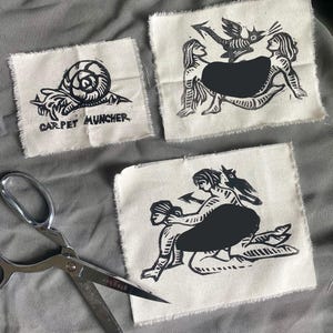 May include: Three fabric patches with black linocut designs. One shows a snail with the text "CARPET MUNCHER." Another depicts two figures with a bird. The third shows two figures in a pose. A pair of silver scissors is also present.