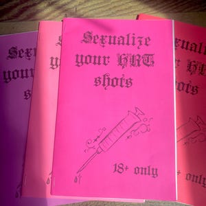 May include: A stack of zines in shades of pink and purple. The cover of the central zine is hot pink and features the text "Sexualize your HRT shots" in gothic lettering, along with a syringe illustration and the text "18+ only."