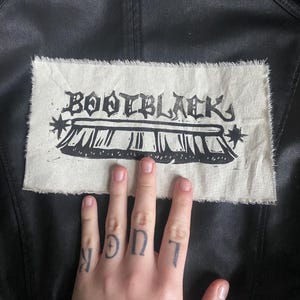 May include: A black jacket with a cream-coloured rectangular patch. The patch has a raw edge and features the text "BOOTBLACK" in a gothic font above a stylized illustration. The jacket is made of a smooth, dark material.
