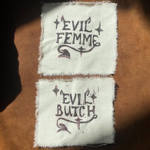 K&ouml;nnte beinhalten: Zwei cremefarbene Stoffflicken mit ausgefransten Kanten. Der obere Flicken tr&auml;gt in dunkelbraunen, stilisierten Buchstaben die Aufschrift "EVIL FEMME". Der untere Flicken tr&auml;gt in &auml;hnlichem Stil die Aufschrift "EVIL BUTCH".