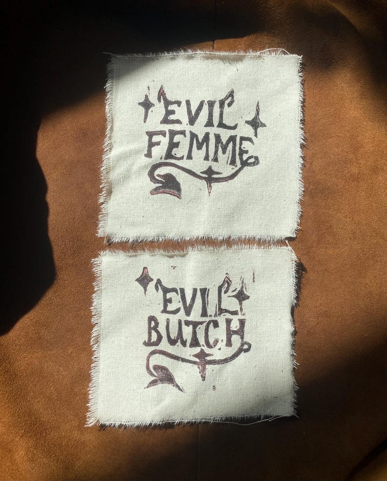 K&ouml;nnte beinhalten: Zwei cremefarbene Stoffflicken mit ausgefransten Kanten. Der obere Flicken tr&auml;gt in dunkelbraunen, stilisierten Buchstaben die Aufschrift "EVIL FEMME". Der untere Flicken tr&auml;gt in &auml;hnlichem Stil die Aufschrift "EVIL BUTCH".