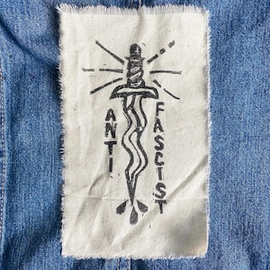 Könnte beinhalten: Jeansjacke mit einem rechteckigen Aufnäher mit einer schwarz-weißen Grafik. Die Grafik zeigt einen stilisierten Dolch, Strahlenlinien und die Worte "ANTI FASCIST" in Großbuchstaben. Der Aufnäher ist auf die Jacke genäht.