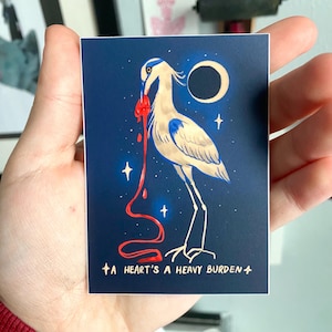 Op de afbeelding: Een rechthoekige kunstdruk met een gestileerde reiger met een rood hart en een bloedachtige stroom tegen een donkerblauwe achtergrond. De print bevat een halve maan, sterren en de tekst "A HEART'S A HEAVY BURDEN."