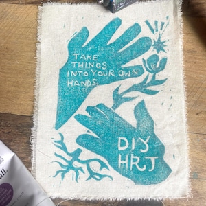 May include: A teal-colored hand-printed design on a canvas square. The text reads "TAKE THINGS INTO YOUR OWN HANDS" and "DIY HRJ". The canvas has a raw, fringed edge. A tube of black ink and a printing roller are in the background.