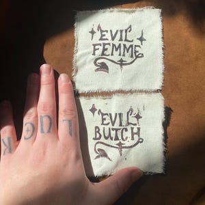 K&ouml;nnte beinhalten: Zwei cremefarbene Stoffflicken mit den W&ouml;rtern "EVIL FEMME" und "EVIL BUTCH" in dunkelbrauner Tinte bedruckt. Die Flicken haben einen rauen, unfertigen Rand. Eine Hand mit dem Tattoo "LUCK" auf den Kn&ouml;cheln ist im Vordergrund.