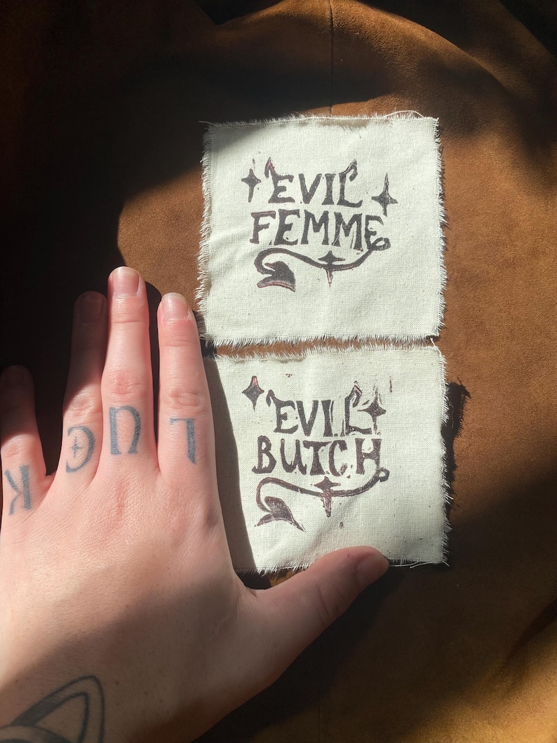 K&ouml;nnte beinhalten: Zwei cremefarbene Stoffflicken mit den W&ouml;rtern "EVIL FEMME" und "EVIL BUTCH" in dunkelbrauner Tinte bedruckt. Die Flicken haben einen rauen, unfertigen Rand. Eine Hand mit dem Tattoo "LUCK" auf den Kn&ouml;cheln ist im Vordergrund.