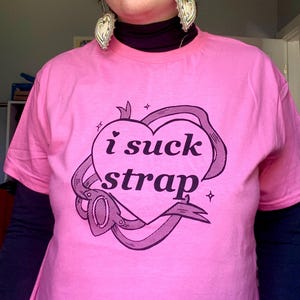 May include: Pink t-shirt with the text "i suck strap" in a heart-shaped design. The graphic is in black and includes a ribbon and stars. The shirt has short sleeves and is worn over a black long-sleeved shirt.
